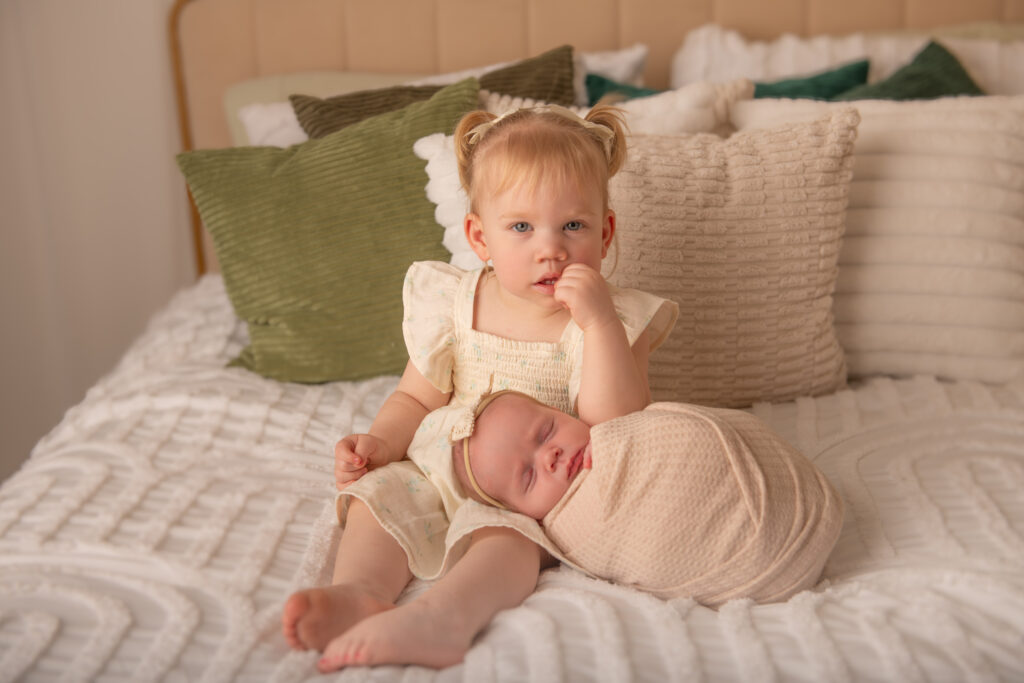 big sister sits with newborn sister on bed wearing white and cream