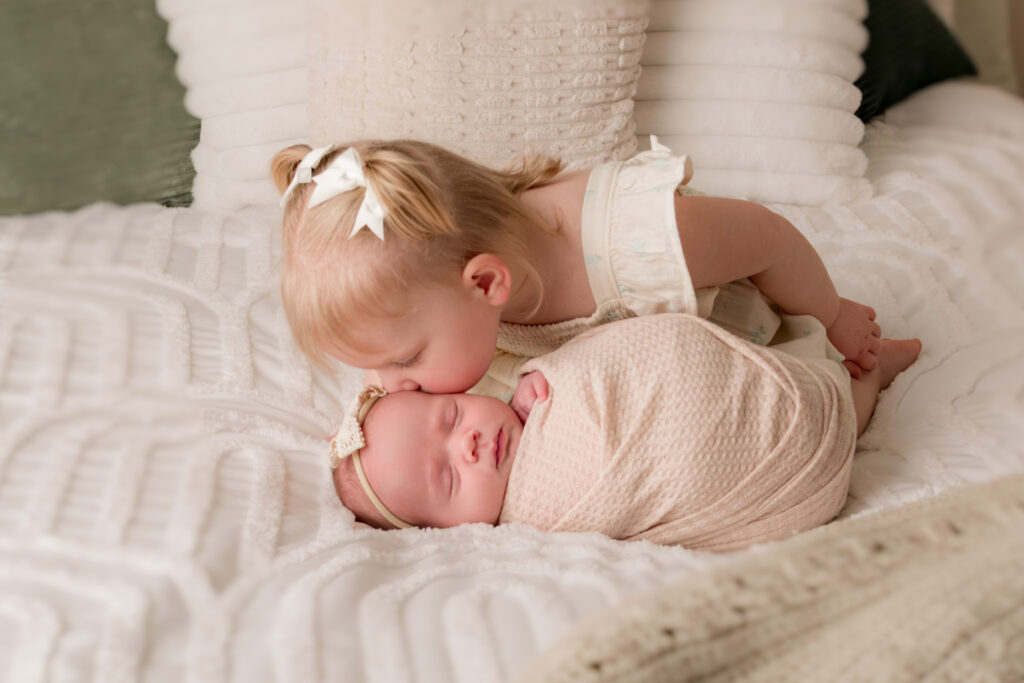 big sister kisses baby sister on bed with white dresses