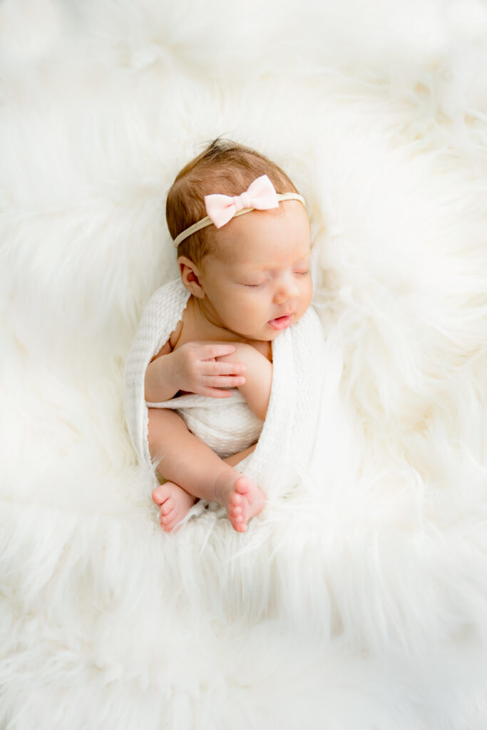 newborn baby girl in white fur with pink bow swaddled in white